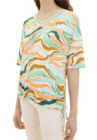 Femme portant un haut ample à col en V avec des motifs abstraits verts, orange et blancs tourbillonnants et un lien latéral réglable, associé à un pantalon de couleur claire.