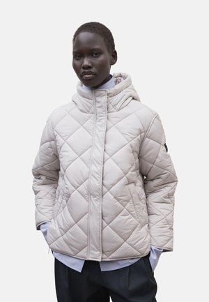 Persona que lleva una chaqueta con capucha acolchada de color beige claro sobre una camisa azul claro y pantalones oscuros, de pie con las manos en los bolsillos.