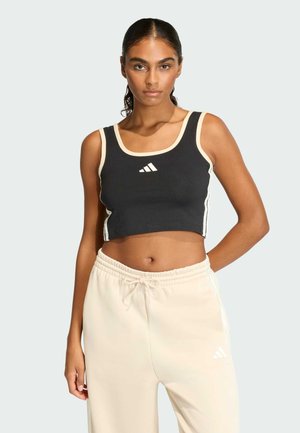 Kobieta z włosami zaczesanymi do tyłu, ubrana w czarny krótki top i kremowe spodnie dresowe, stojąca na tle jednolitego jasnego tła.