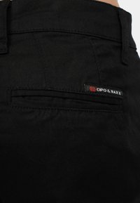 Un pantalon noir en coton présente une poche arrière avec une étiquette de marque rouge et blanche. Le tissu a une texture lisse et durable.