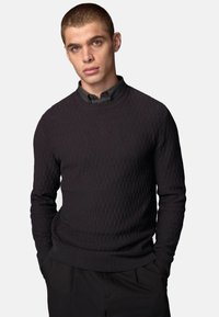 Mørk tekstureret sweater med ribbet mønster, rund halsudskæring og lange ærmer, parret med en skjorte med krave nedenunder.