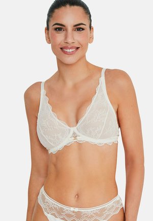 Vrouw draagt een witte kanten beha en bijpassende kanten slip, glimlachend met haar naar achteren gekamd tegen een effen achtergrond.