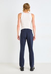 Pantalons bleu marine à coupe ajustée, avec des poches arrière et une texture lisse. Portés avec un débardeur blanc sans manches et des bottes noires.