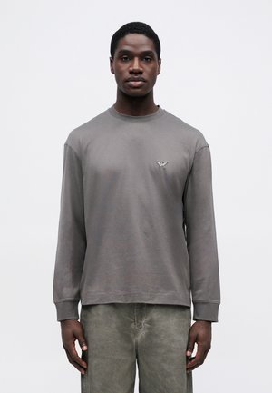 Graues Langarm-T-Shirt mit Rundhalsausschnitt und Logodetail auf der linken Brust; weicher, glatter Stoff, lockere Passform, kombiniert mit hellgrauen Hosen.