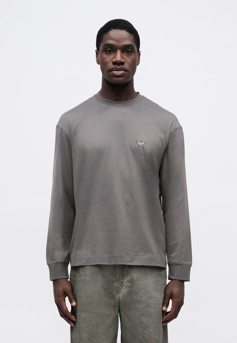 Pelēks garām piedurknēm T-krekls ar apaļo kakla izgriezumu un logo detaļu kreisajā krūtī; mīksta, gluda auduma, brīvas formas, kombinēts ar gaiši pelēkām biksēm.