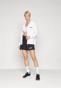 Fehér cipzáras kapucnis felső, fekete sportnadrág elasztikus derékkal, fehér crop top, fekete sportcipő és fehér zokni fekete részletekkel.