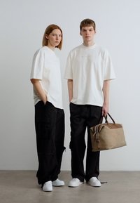 Δύο άτομα φορούν φαρδιές λευκές μπλούζες και μαύρο παντελόνι. Το ένα κρατά μια καφέ τσάντα με δερμάτινες λεπτομέρειες. Λευκά αθλητικά παπούτσια ολοκληρώνουν την εμφάνιση.