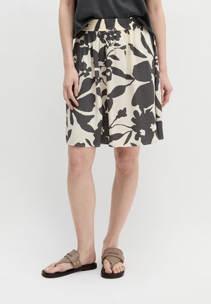 Femme portant une jupe beige et noire à fleurs jusqu'aux genoux, un haut noir et des sandales plates beiges, debout sur fond blanc.