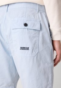 Calções azul claros feitos de um tecido com textura amarrotada. Apresentam um bolso traseiro com aba, botão preto e bordado "BARBOUR INTERNATIONAL".