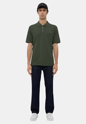 Polo shirt verde scuro con colletto e tre bottoni, abbinato a jeans scuri e sneakers bianche, indossato con un cappellino nero.