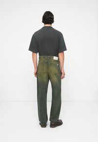 Tricou oversized de culoare gri închis și blugi verzi măslin cu efect de gradient, având o curea și buzunare la spate. Pantofi maro completează aspectul.