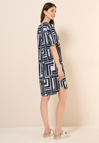 Marineblaues und cremefarbenes Kurzarmkleid mit geometrischen Drucken, lockerer Passform, seitlichen Schlitzen und weichem Material. Kombiniert mit metallischen Slippern.