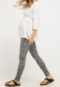 Bílá blůza s páskem a tříčtvrtečními rukávy, kombinovaná se šedými legínami s peříčkovým vzorem a nařasenými detaily, nošená s černými sandály.