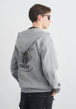 Mladý muž v sivom mikine s kapucňou s čiernou grafikou a textom na chrbte, v čiernych roztrhaných džínsoch, so slnečnými okuliarmi a strieborným náramkom, stojaci zboku.