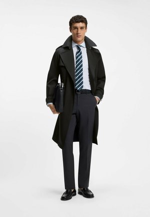 Mand i sort trenchcoat, hvid skjorte, stribet slips, mørke bukser, der holder en sort mappe, står mod en lys ensfarvet baggrund.