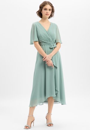 Junge Frau trägt ein hellgrünes Wickelkleid mit flatternden Ärmeln und hochhackigen Sandalen mit Knöchelriemen, steht vor weißem Hintergrund.