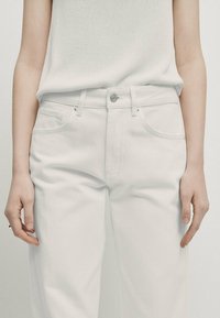 White denim jeans featuring a fitted style, silver button closure, belt loops, and four pockets. The fabric is smooth with minimal texture.