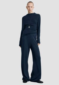 Bluză tricotată bleumarin cu mâneci lungi și un decolteu unic, asortată cu pantaloni largi bleumarin dintr-un material moale. Colier cu pandantiv argintiu.