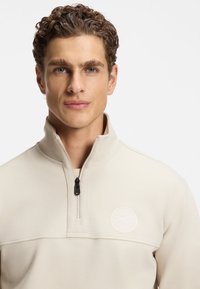 Beżowy pulower z zamkiem do ćwiartki, wykonany z teksturowanej tkaniny, z małym logo na piersi i czarnym zamkiem błyskawicznym. Dopasowany krój z wysokim kołnierzem.