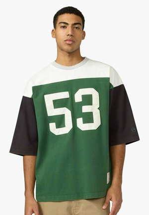 Giovane uomo che indossa una maglia larga verde, nera e bianca con il numero 53 in grande stampa bianca sul petto.