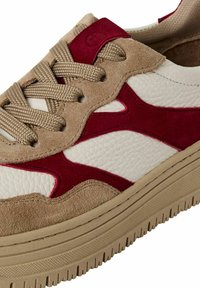 Zapatillas beige y blancas con acentos de gamuza roja, cuero texturizado y cordones planos beige. Presentan una suela gruesa y estriada de color marrón claro.