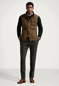 Άνδρας που φοράει καφέ ραμμένο γιλέκο πάνω από σκουροπράσινο πουλόβερ και μπλε πουκάμισο, γκρι παντελόνι και καφέ δερμάτινα παπούτσια, στέκεται με τα χέρια στις τσέπες.