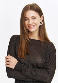 Czarna bluzka z długim rękawem o teksturowanym, błyszczącym wykończeniu, charakteryzująca się luźnym krojem i okrągłym dekoltem. Modelka ma na sobie perłowe kolczyki w kształcie kół.