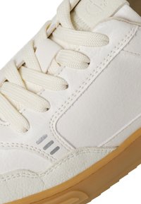 Vit sneaker med ovandel i slät läder, texturerade snören och en sula i gummigummi. Har silveraccent och förstärkta sömmar.