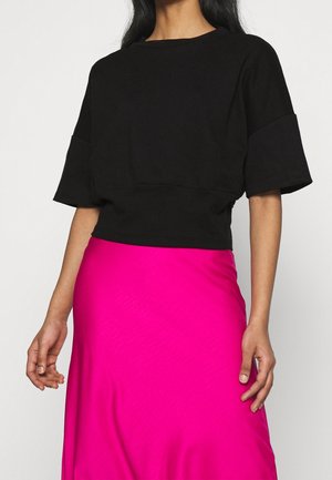 Person trägt ein schwarzes, locker sitzendes Kurzarmoberteil und einen leuchtend pinken, hoch taillierten Rock vor einem schlichten hellen Hintergrund.