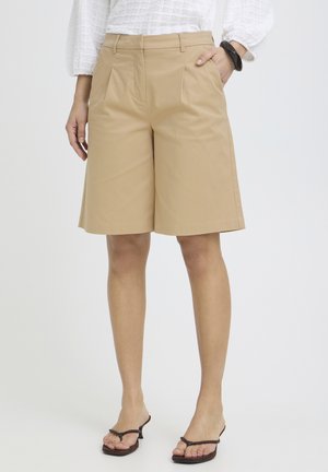 Nő bézs, térdig érő, szabott rövidnadrágban, fehér texturált blúzban, fekete karkötővel és fekete, nyitott orrú, magas sarkú szandálban, egyik kezét zsebre téve áll.