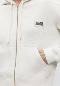 Personne portant un sweat à capuche beige clair avec zip, poche avant et petit patch rectangulaire avec logo sur la poitrine. La main est dans la poche.