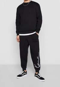 Persona vistiendo sudadera negra, pantalones de chándal negros con escritura blanca en la pierna izquierda, camiseta blanca, zapatillas de deporte negras y blancas, y collar de cadena plateada.