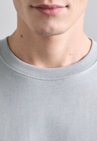 Světle šedá bavlněná mikina s kulatým výstřihem, která má vroubkovaný límec a hladkou strukturu, zaměřená na detaily výstřihu a oblasti brady.