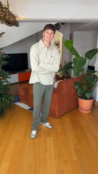 Light gray long-sleeve shirt with a collar, olive green ribbed pants, and shiny silver sneakers. Indoor setting with a plant and couch.
