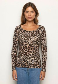 Frau in einem langärmligen Leopardenmuster-Oberteil und blauen Jeans, die vor einem schlichten hellen Hintergrund steht und nach vorne schaut.