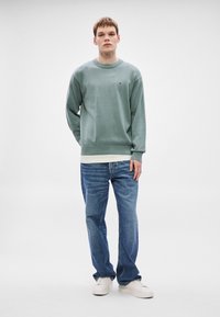 Πράσινο φούτερ με στρογγυλή λαιμόκοψη και ριμπ μανσέτες, φορεμένο πάνω από ένα λευκό πουκάμισο. Συνδυάζεται με μπλε τζιν και λευκά αθλητικά παπούτσια. Απλός σχεδιασμός.