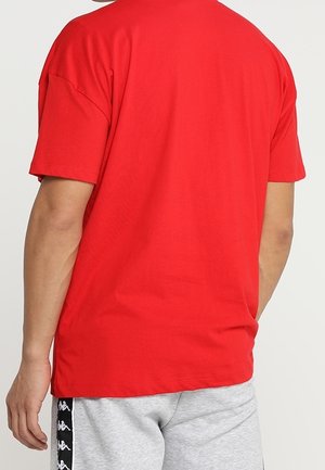 Person iført en ensfarvet rød kortærmet skjorte og lysegrå shorts med sorte og hvide logostriber, set bagfra.