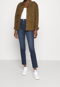 Olijfgroene jacket met grote knoppen en twee voorzakken, gecombineerd met donkerblauwe jeans en witte sneakers. Eenvoudig, gestructureerd ontwerp.