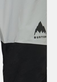 Kurtka Burton z szarym górnym segmentem, czarnym dolnym segmentem, gładkim materiałem i czarnym logo góry na piersi.