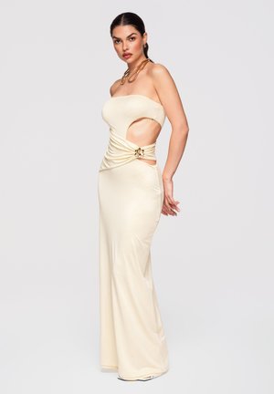 Kobieta w kremowej, bezramkowej sukni wieczorowej z bocznymi wycięciami i skręconym detalem, nosząca złoty naszyjnik i kolczyki na jednolitym tle.