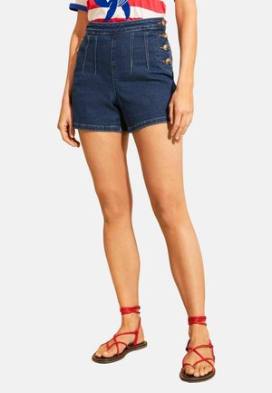 Naine, kes kannab kõrge vöökohaga tumedat sinist teksashortsit külgsete nööpidega ja punaseid krõpssandaale, seisab ühevärvilise tausta ees.