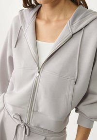 Hellgraue Zip-Hoodie mit einer Kordelzugkapuze, Fronttaschen und elastischen Bündchen. Weicher Stoff mit glatter Textur.