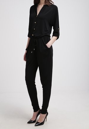 Kobieta ubrana w czarny kombinezon na guziki z talią ze sznurkiem i rękawami do łokcia, w połączeniu z czarnymi szpiczastymi wysokimi obcasami.