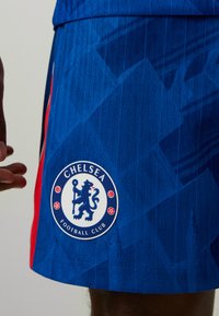 Blauwe voetbalkorte broek met een gestructureerd ontwerp, rode zijkanten en een ronde Chelsea Football Club-logo op de linkeronderkant.