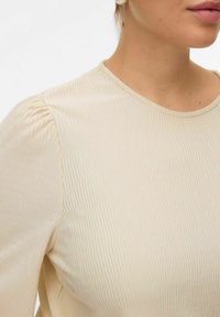 Kreemivärviline pluus ribade tekstuuriga, ümar kaelus ja puffidega varrukad, millel on sile kangas.