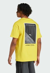 Gul t-skjorte i bomull med et bakre trykk av en månescene. Detaljer inkluderer teksten "SE OPP FRA JORDEN TIL ROMMET" og merkevarelogoer.