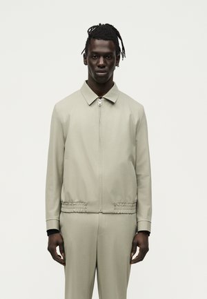 Homme aux cheveux tressés portant une veste beige zippée avec une ceinture élastique et un pantalon beige assorti sur un fond clair uni.