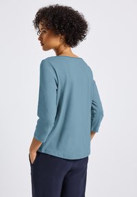 Maglietta a maniche lunghe in morbido e leggero tessuto teal. Presenta un collo rotondo e una vestibilità rilassata. La vista posteriore mostra un design casual e una leggera caduta.