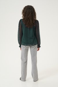 Blusa in pizzo verde scuro con motivi geometrici, maniche lunghe trasparenti e polsini arricciati. Abbinata a jeans a gamba larga grigio chiaro.