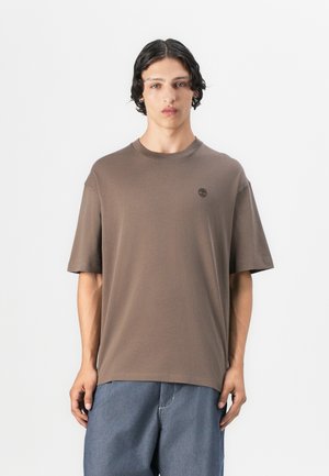 Brown short-sleeve t-shirt made of soft fabric, featuring a crew neck and a small logo on the chest. Paired with loose blue denim pants.
